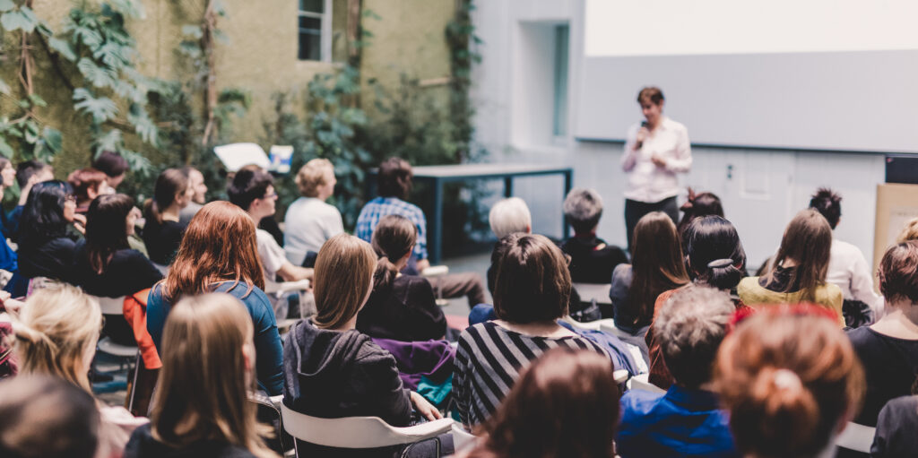 Female speaker giving presentation in lecture hall at university workshop. Audience in the conference hall. Rear view of unrecognized participant in audience. Scientific conference event.